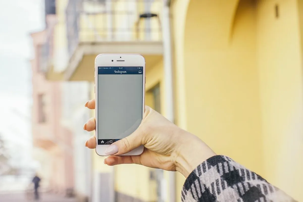 Main tenant un smartphone ouvert sur un fil d'actualité de réseau social, dans une rue aux façades colorées